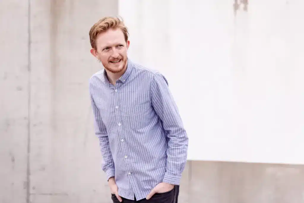 A man with light brown hair and a short beard, wearing a blue striped shirt and dark trousers, stands outdoors with his hands in his pockets, smiling and looking to the side.