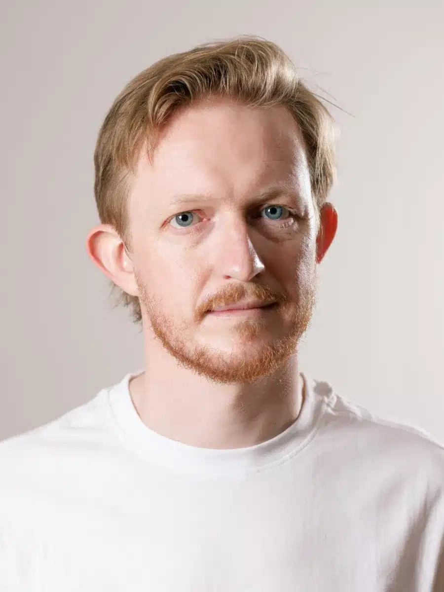 A man with short blond hair, a light beard, and blue eyes looks at the camera. He is wearing a plain white T-shirt and is posed against a neutral light background.