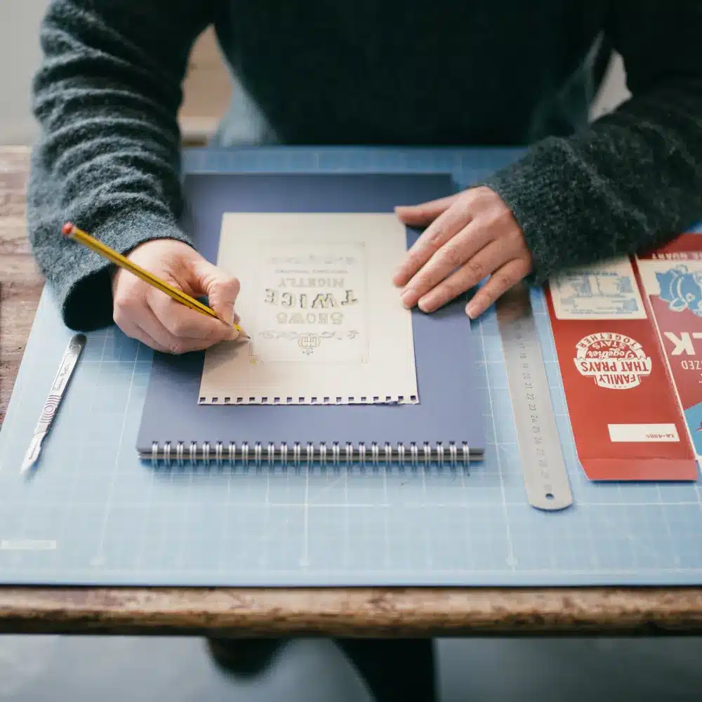 A person sketches on a notepad with a pencil at a desk, surrounded by a ruler, craft knife, and colourful design samples on a cutting mat.