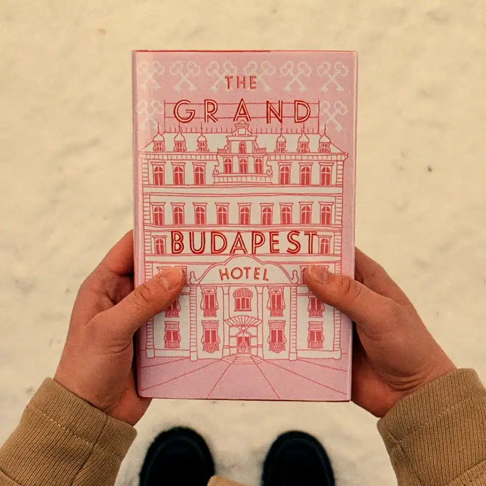Two hands hold a pink book titled "The Grand Budapest Hotel" against a light background, with a drawing of a grand building on the cover and only the person's lower arms and shoes visible.