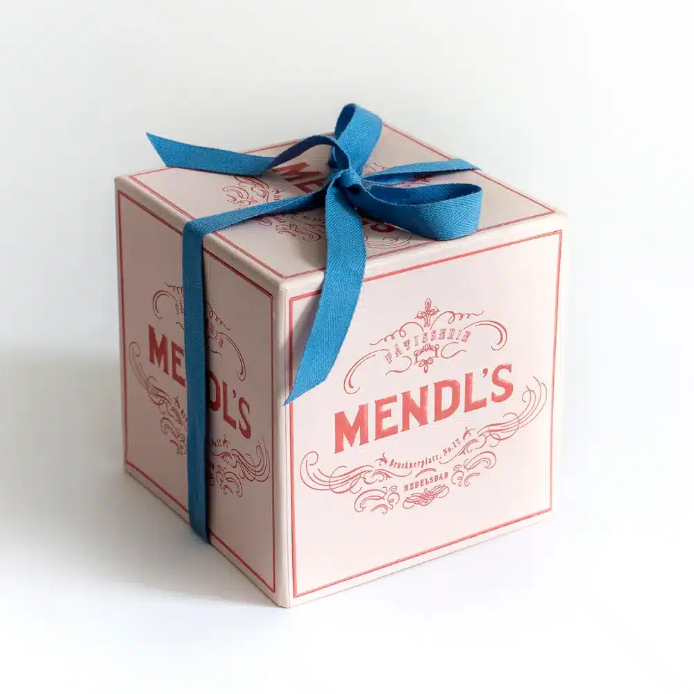 A pink box labelled "Mendl's" with decorative text, tied with a blue ribbon, sits on a white background.