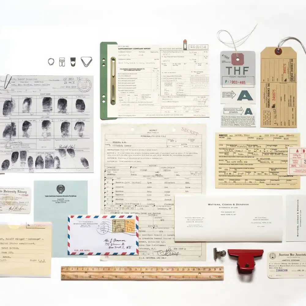 A flat lay of vintage documents, ID cards, fingerprint record, envelopes, tags, a red wax seal, and a wooden ruler arranged neatly on a white background.