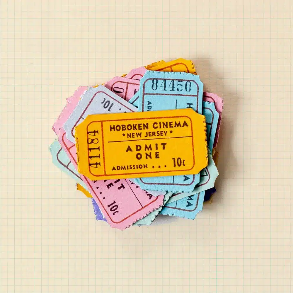 A stack of colourful vintage admit-one tickets, with the top yellow ticket reading "Hoboken Cinema, New Jersey, Admit One, Admission 10p," on a squared paper background.
