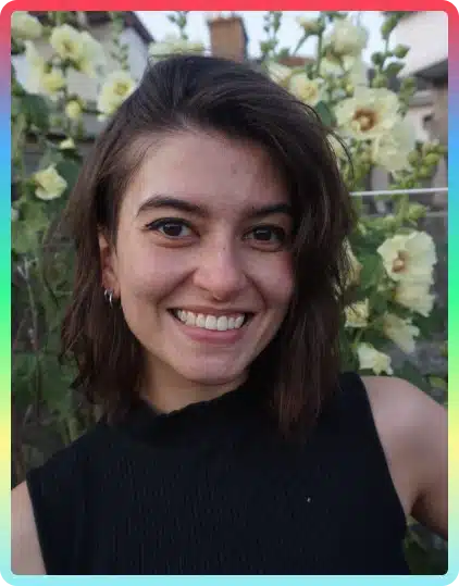 A young woman with shoulder-length brown hair smiles in front of tall yellow flowers at the Kilkenny Animated Festival. She wears a sleeveless black top and silver hoop earrings. The background shows blurred greenery and a building.