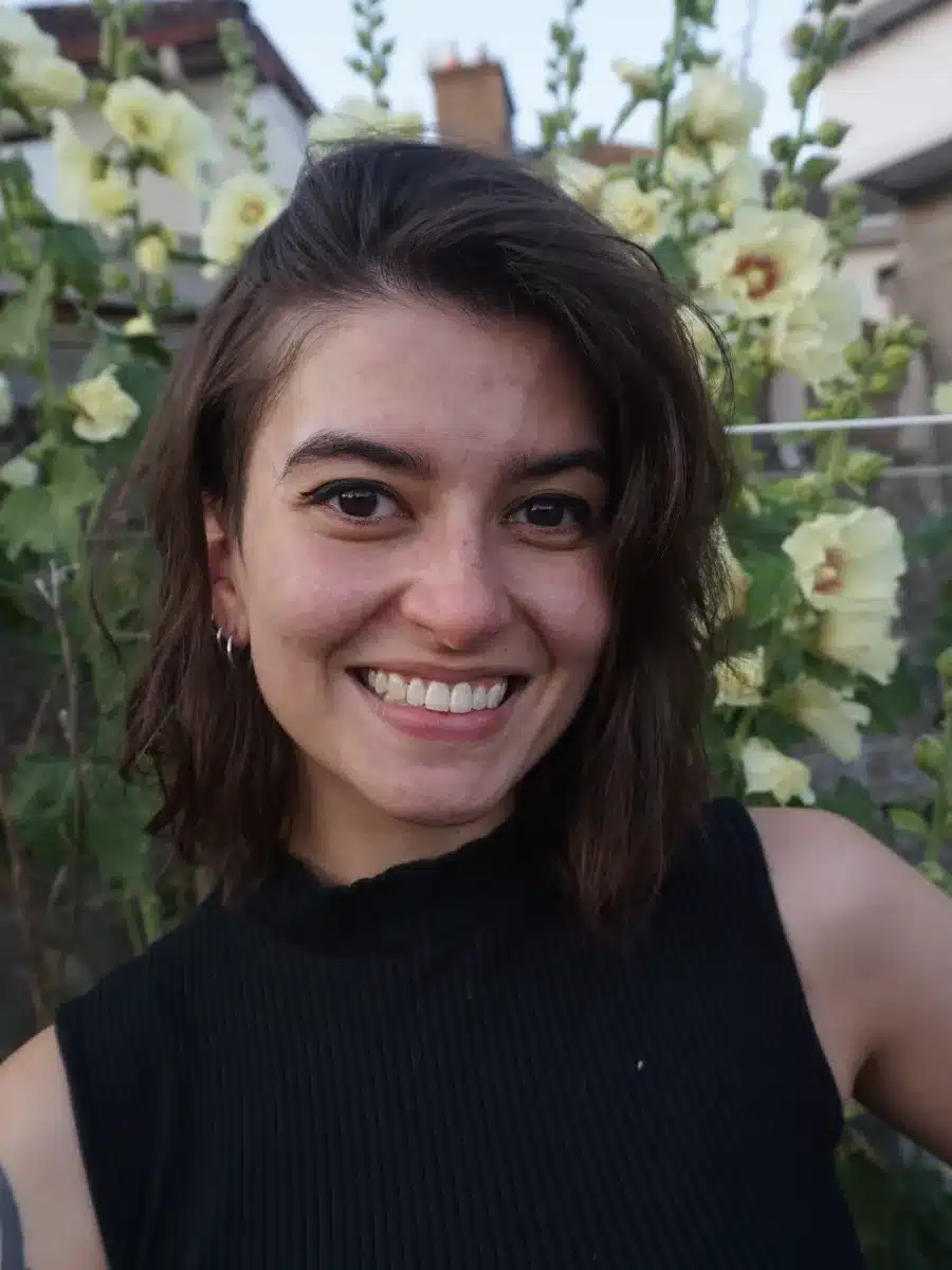 A young woman with shoulder-length brown hair smiles outdoors in front of tall green plants with pale yellow flowers. She is wearing a sleeveless black top.