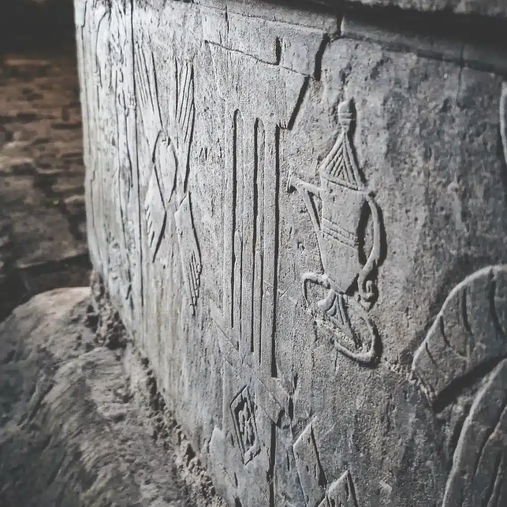 Close-up of ancient stone carvings featuring detailed symbols and patterns, including a vase-like object and geometric forms, on a weathered wall with a rough, textured surface.