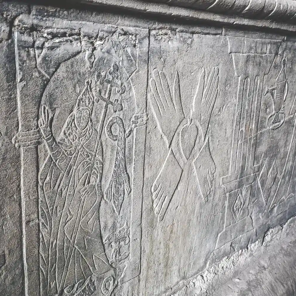Stone carving shows a robed figure holding a crosier, crossed arms with open hands, columns, and a hanging lamp, all etched into a weathered stone wall.