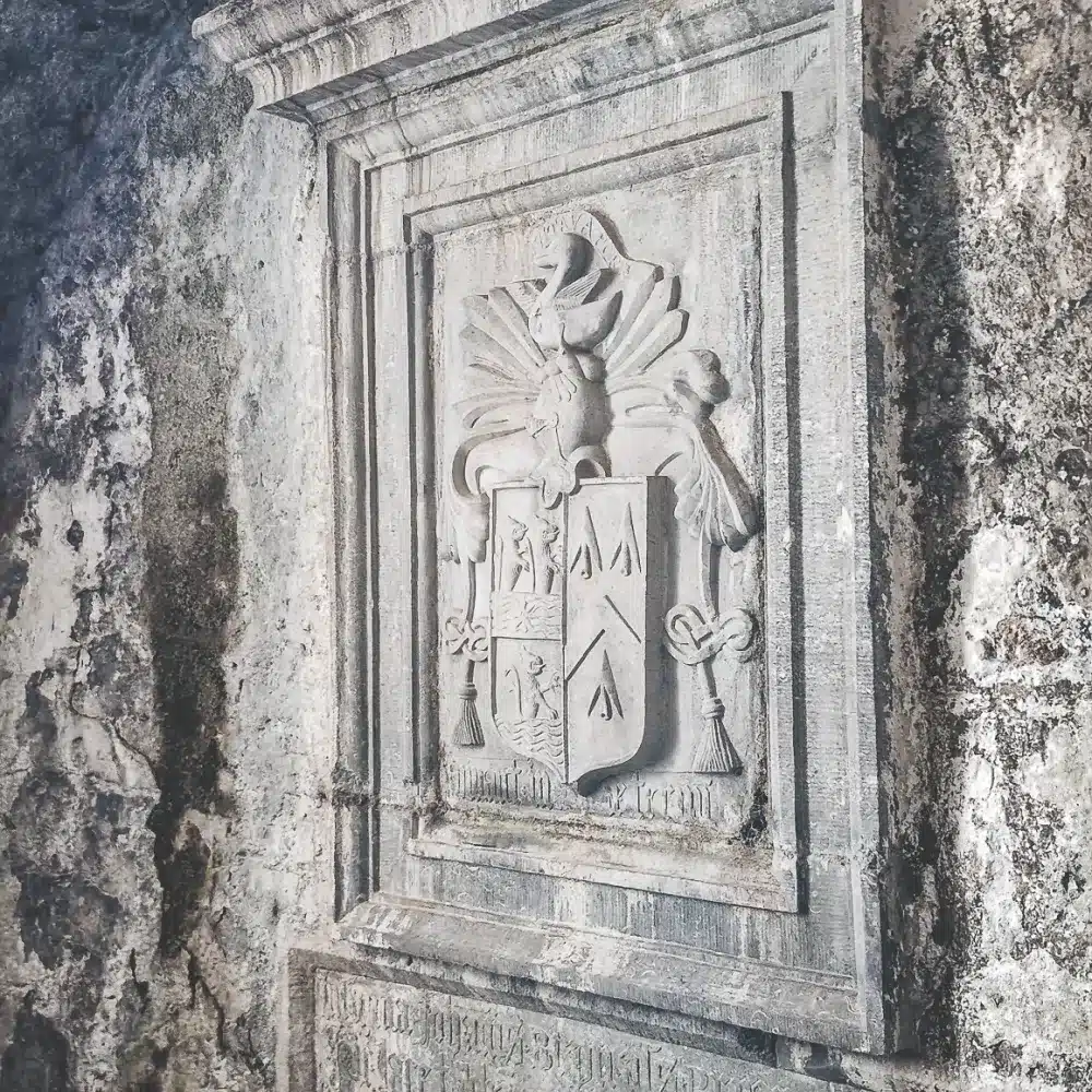 Stone relief of a heraldic coat of arms with a shield, decorative flourishes, and what appears to be a bird or dragon atop, mounted on a weathered wall.