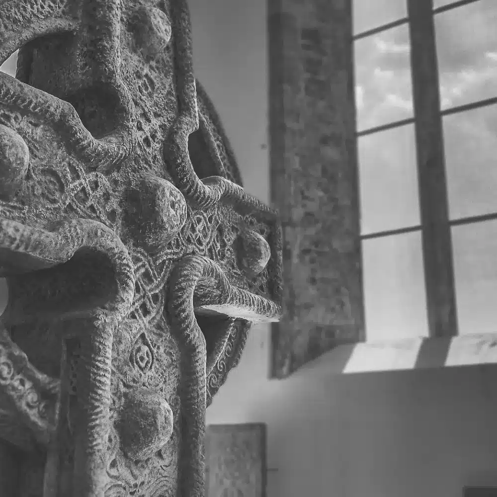 A close-up of a carved Celtic cross with intricate patterns, positioned indoors near tall windows that let in natural light. The photo is in black and white.