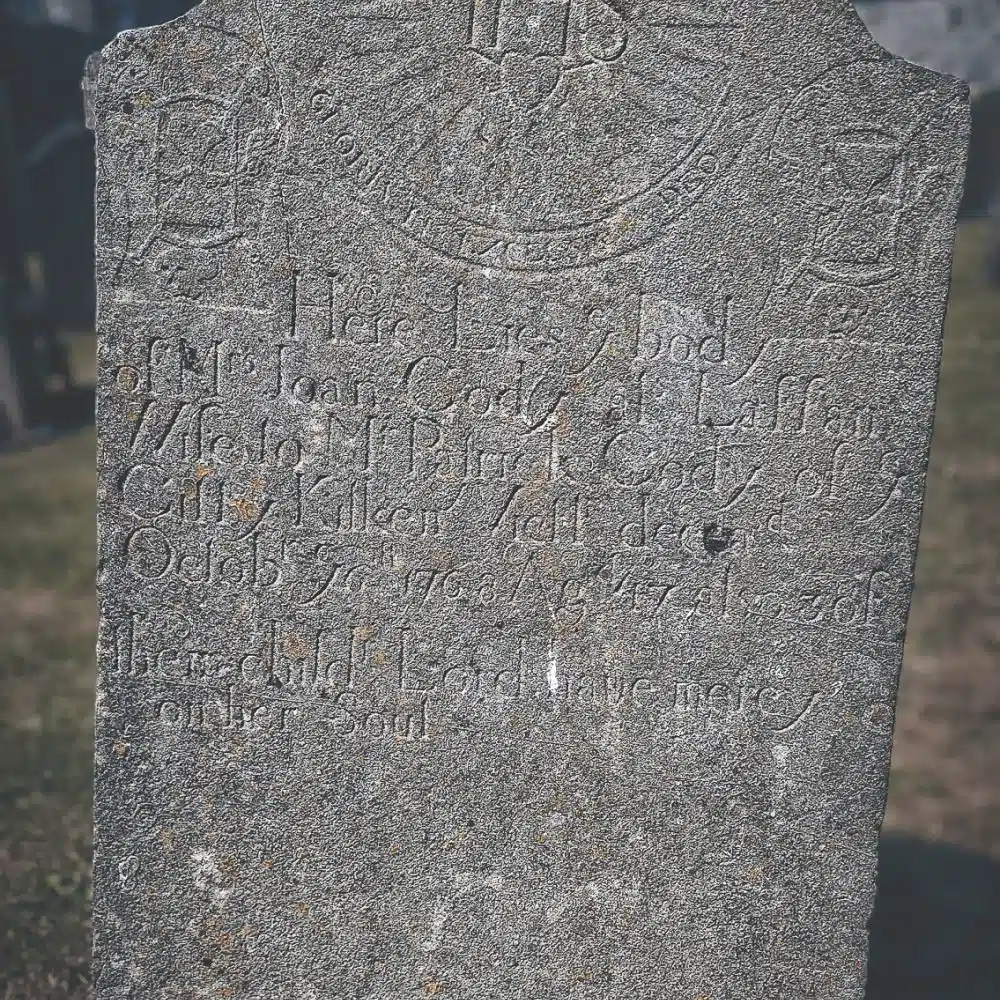 A weathered grey gravestone with engraved text and faint carvings, including a winged face and decorative patterns, stands on a grassy area in a cemetery. The text is partially eroded and difficult to read.