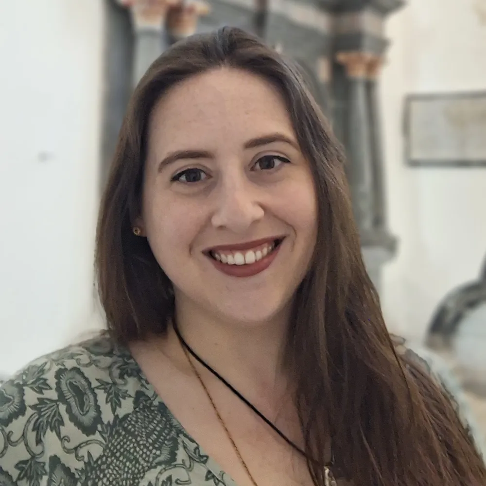 A woman with long brown hair smiles at the camera, wearing a green patterned top and a black necklace. The background shows blurred architectural details and pale walls.