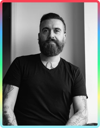 A bearded man with tattoos, wearing a black t-shirt, sits by a window. The image is in black and white with a colourful border inspired by the vibrant energy of the Kilkenny Animated Festival.