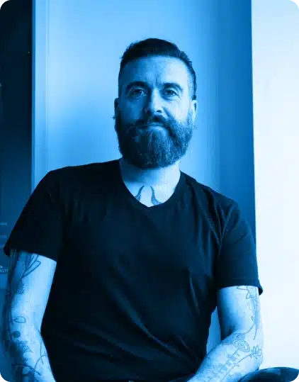 A bearded man with tattoos on his arms, wearing a black T-shirt, sits near a window at the Kilkenny Animated Festival. The image has a blue colour filter.