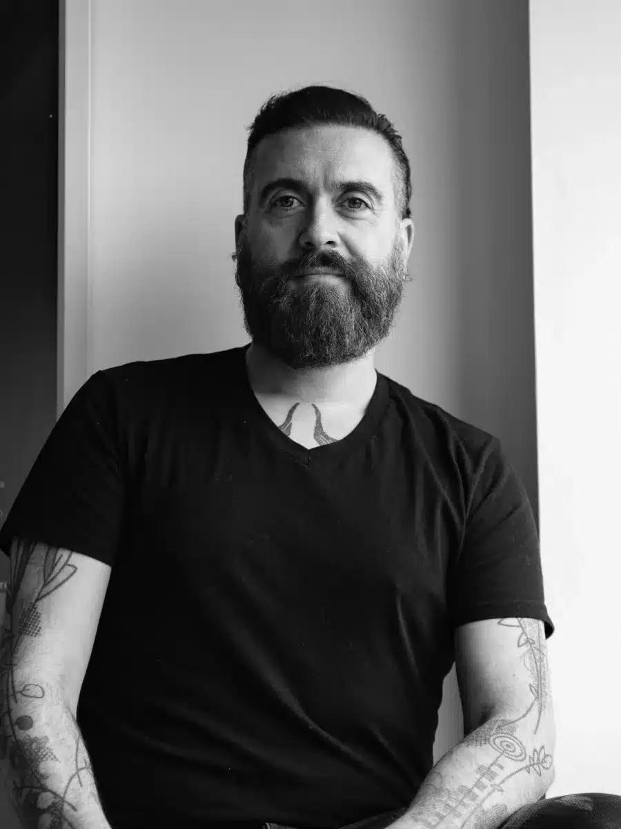 A bearded man with tattoos and a black T-shirt sits by a window, looking at the camera with a calm expression. The photo is in black and white.