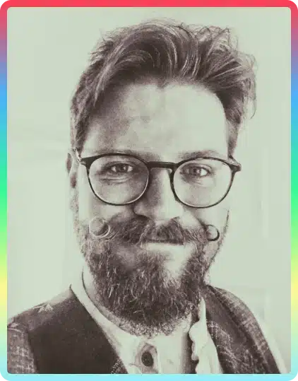 A bearded person with glasses and a curled moustache smiles at the camera. The photo is black and white with a border in rainbow colours, capturing the vibrant spirit of the Kilkenny Animated Festival.