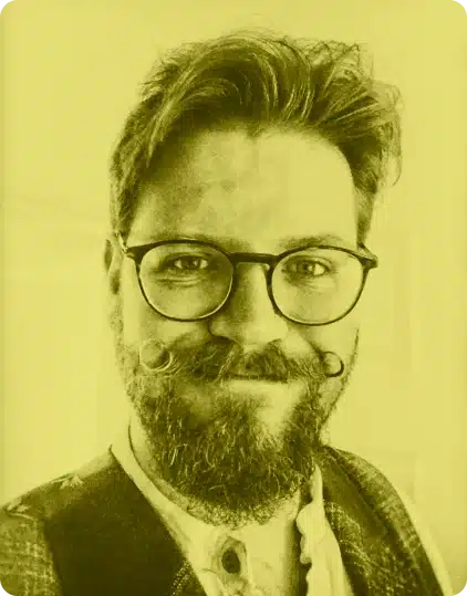 A person with glasses, a curled moustache, and a beard smiles at the camera at the Kilkenny Animated Festival. They wear a patterned waistcoat over a collared shirt. The photo has a yellow tint.