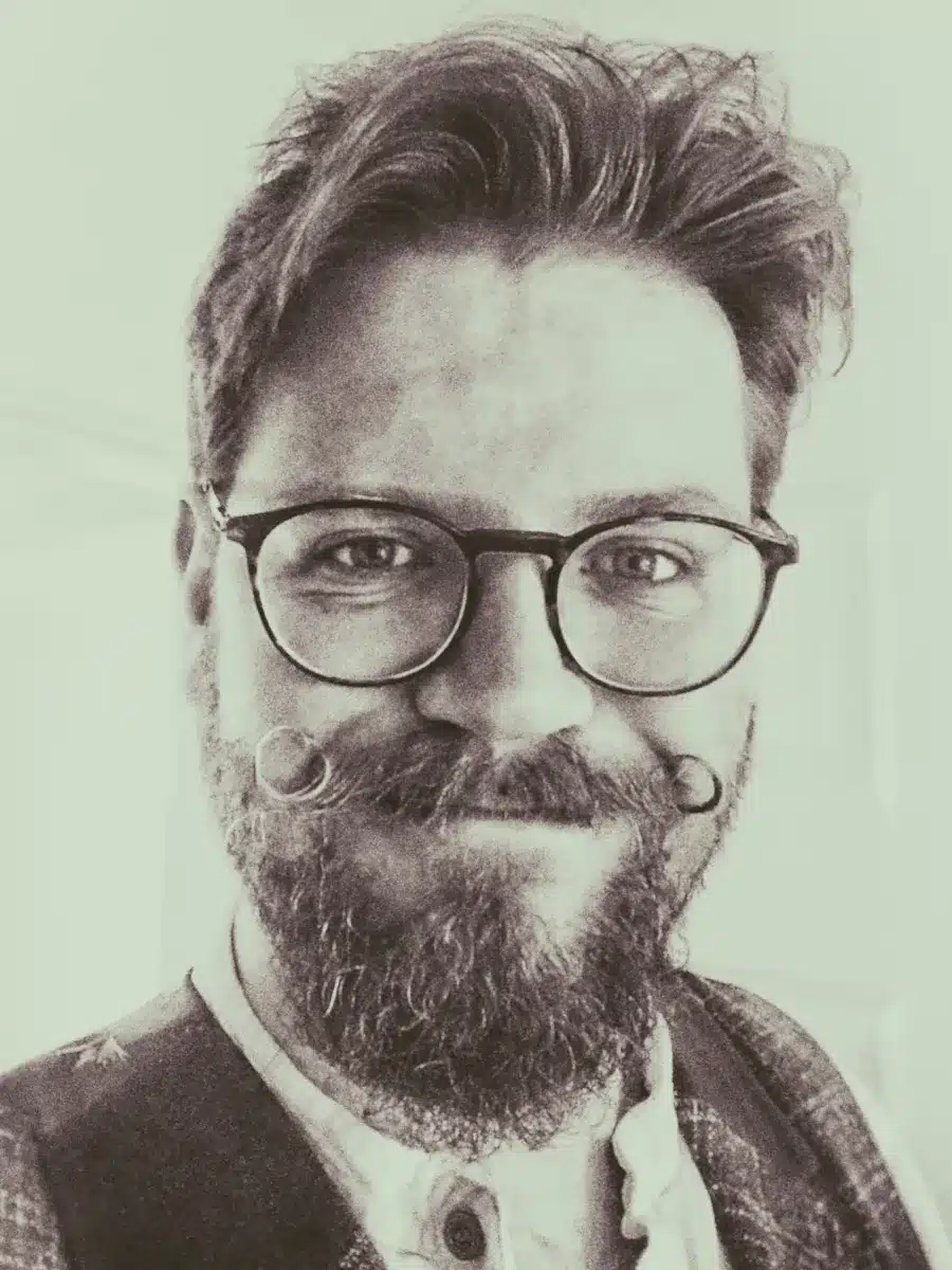 A bearded person with round glasses and a curled moustache smiles at the camera. They have wavy hair, wear a button-up shirt, and a jacket. The photo is in black and white.
