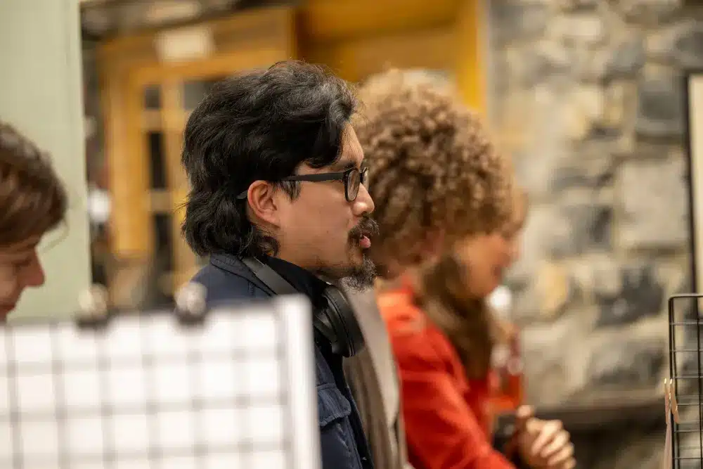 A man with glasses and headphones around his neck is talking, with two people blurred in the background against a stone wall.