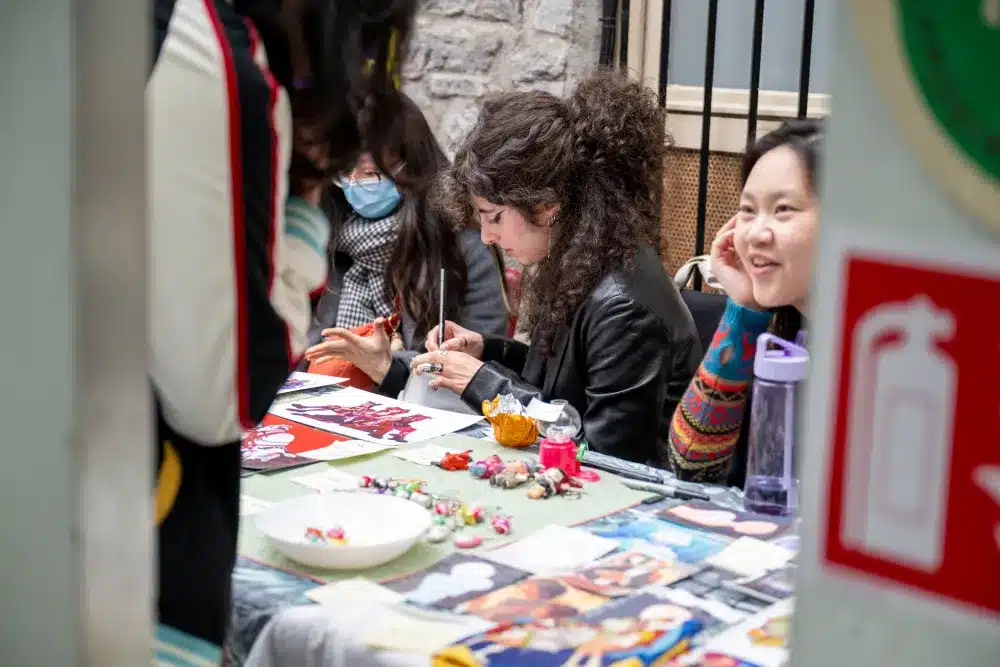 Three women sit at a table covered with colourful art prints and crafts. One paints a small object, another smiles at someone off-camera. A fire extinguisher sign is visible in the foreground.