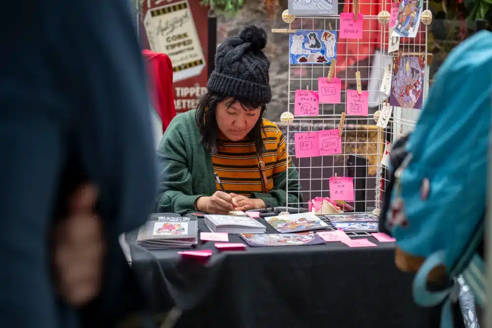 A person in a striped shirt and black hat sits at a table drawing, surrounded by art prints and pink price labels at a market or convention stall.