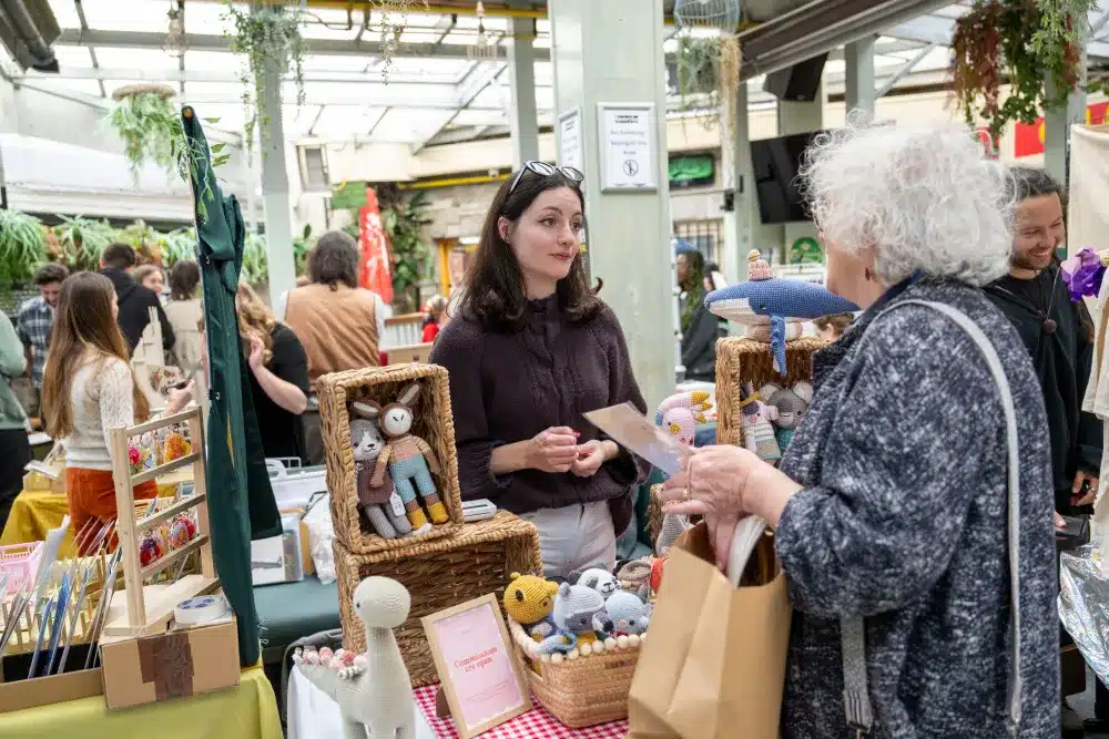 Two women talk at a craft market stall displaying handmade toys and crafts. Other shoppers browse in the background amid a bright, indoor setting with green plants and natural light.