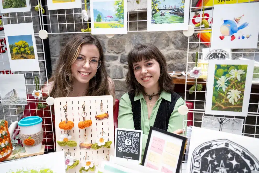 Two smiling women sit at a craft stall displaying art prints, crochet items, and a framed QR code. Colourful decor and artwork hang on a wire grid behind them. The background features a stone wall.