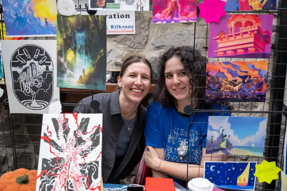 Two people smile at a colourful art stall, surrounded by vibrant prints and paintings displayed on a wire rack. A stone wall is in the background, and a coffee cup is visible on the table in front of them.