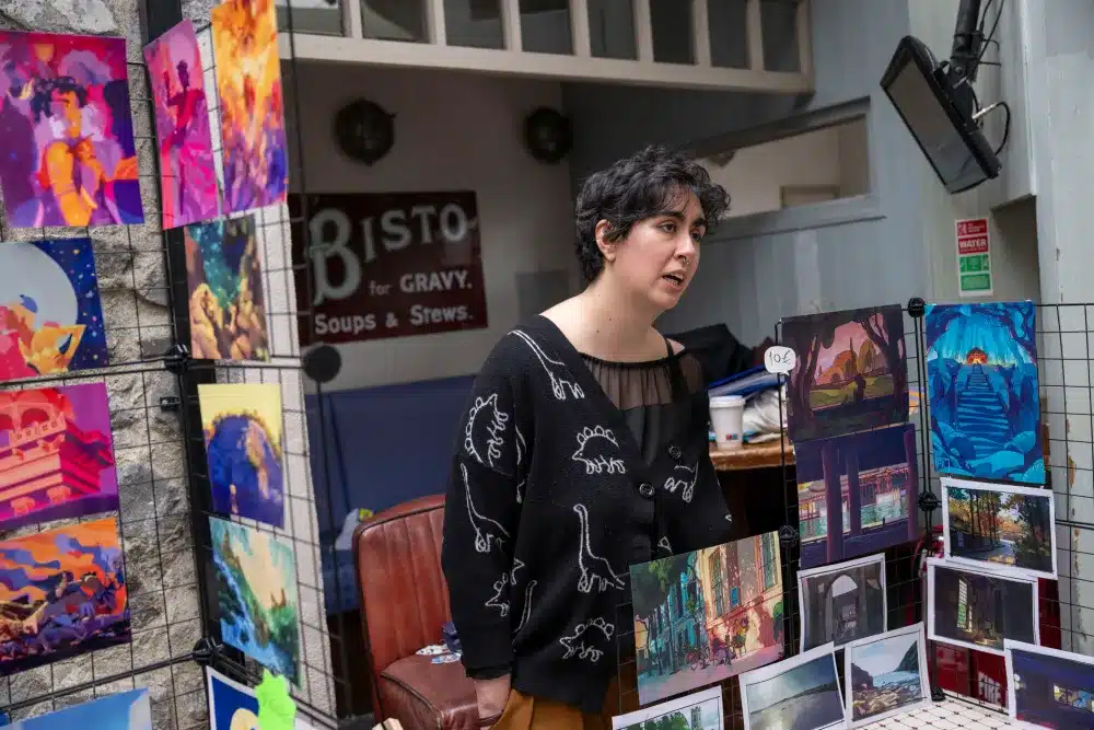 A person stands behind a display of colourful art prints at an indoor market. They wear a black jumper with dinosaur designs and appear to be talking to someone out of frame. Artworks feature vibrant landscapes and cityscapes.