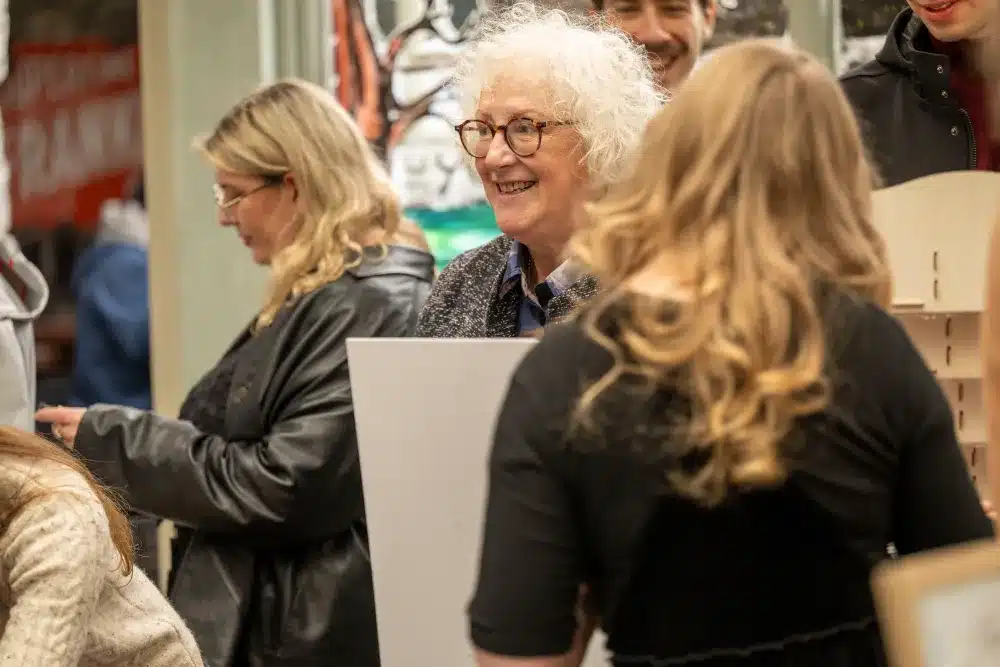 A smiling older woman with curly white hair and glasses stands among a group of people indoors, holding a white card. Others around her are talking and interacting, with blurred artwork visible in the background.