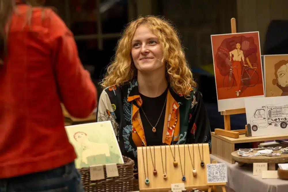 A woman with curly blonde hair smiles whilst sitting at a craft stall displaying jewellery and art prints. Another person, seen from behind, is talking to her. The table shows colourful necklaces, drawings, and postcards.