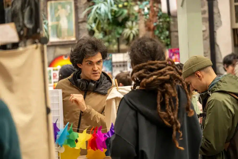 A man with headphones round his neck talks to a woman with dreadlocks at a colourful indoor market, whilst another person browses nearby.