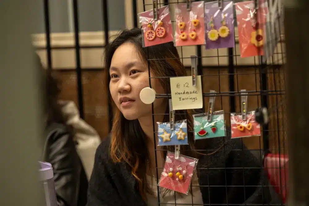 A woman sits behind a display of handmade earrings at a market stall. The earrings are colourful, hanging on a wire grid, with a sign reading "€12 Handmade Earrings.