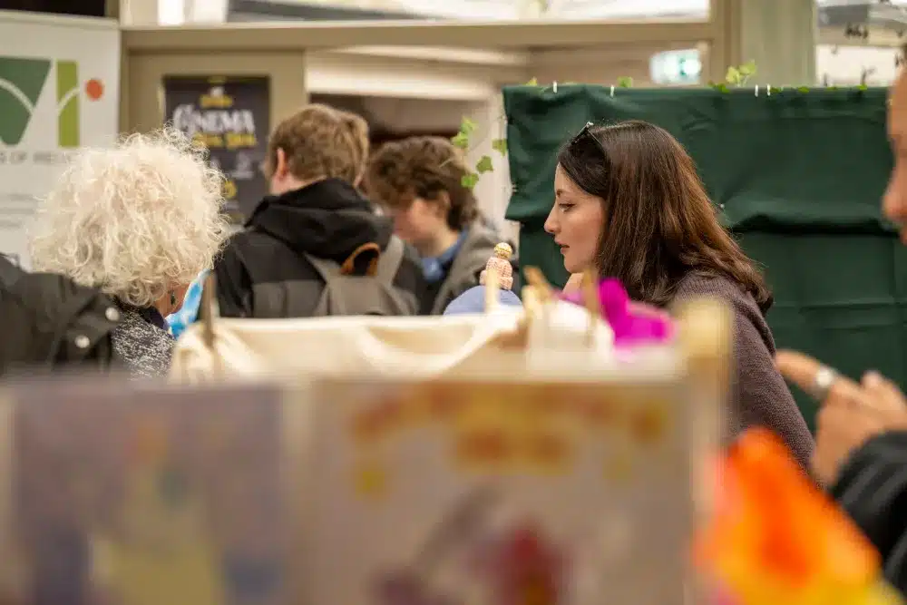 A woman eats ice cream in a crowded indoor space, surrounded by people and colourful decorations. The scene appears lively and social.