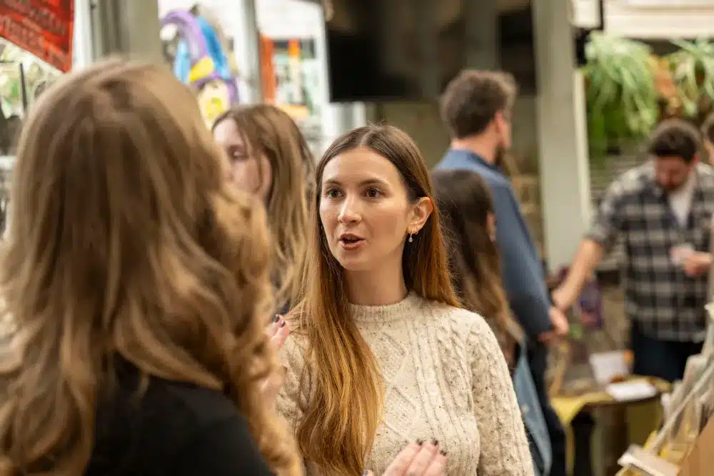 A woman with long brown hair in a cream jumper speaks to another woman at a busy indoor event. People are gathered in the background, some browsing tables and displays.