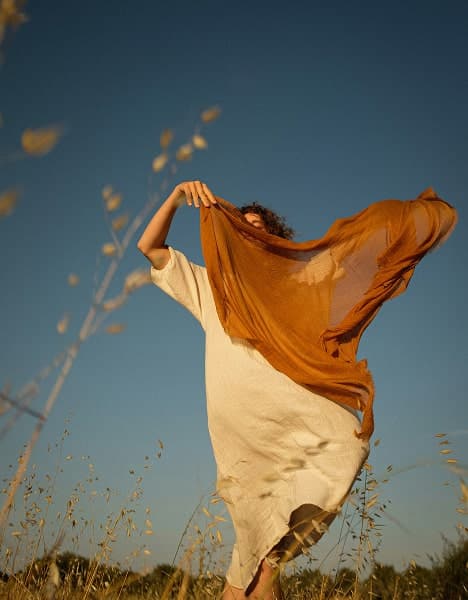 A person in a light dress stands in a field, holding up a flowing orange scarf against a clear blue sky, with tall grass in the foreground.