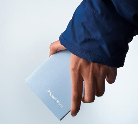 A person wearing a blue jacket holds a small, light blue notebook labelled "Pocket Note" against a plain background.