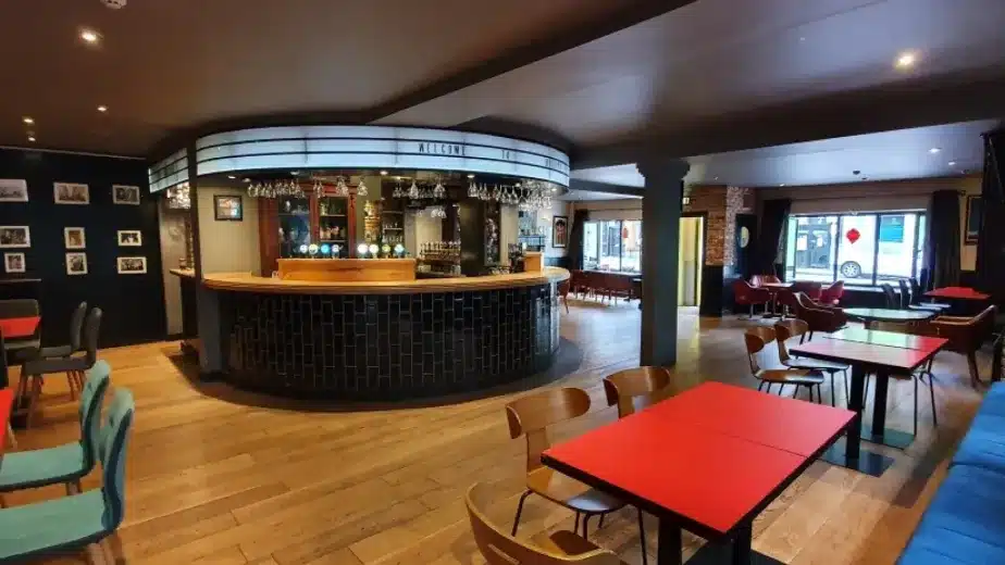 Modern bar with wooden floors, red tables, mixed seating, brick walls, and a curved counter. Shelves with bottles and glassware are behind the bar. Large windows provide natural light.