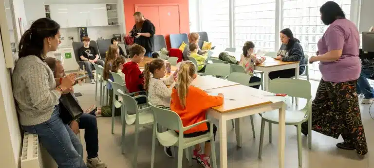 Children sit at tables in a bright classroom, engaged in activities. Two adults stand nearby, while another adult sits on the left. Art supplies and exercise books are on the tables. The scene is lively and collaborative.