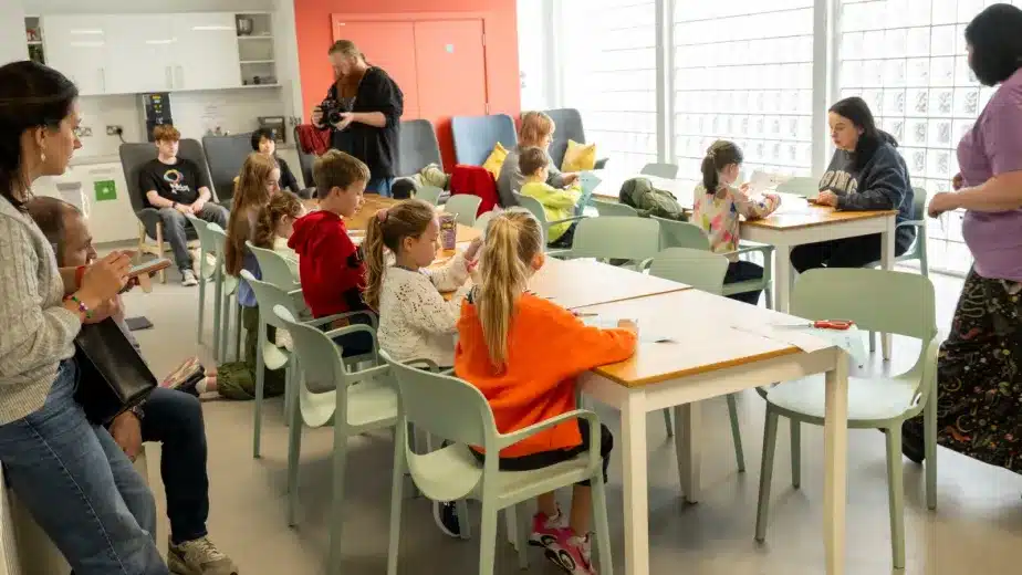 Children sit at tables in a bright classroom, engaged in activities. Two adults stand nearby, while another adult sits on the left. Art supplies and exercise books are on the tables. The scene is lively and collaborative.