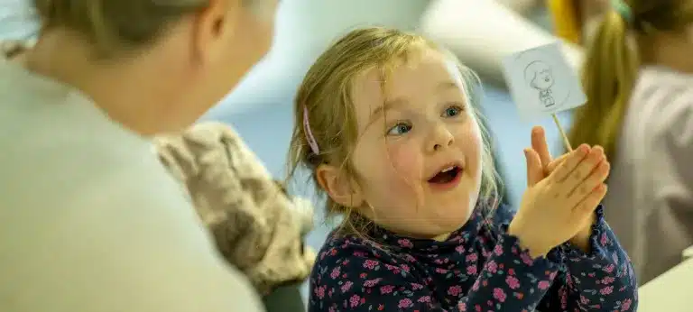 A young girl with blonde hair, wearing a floral shirt, smiles excitedly whilst holding a small stick with a drawing on it, as an adult beside her looks on.
