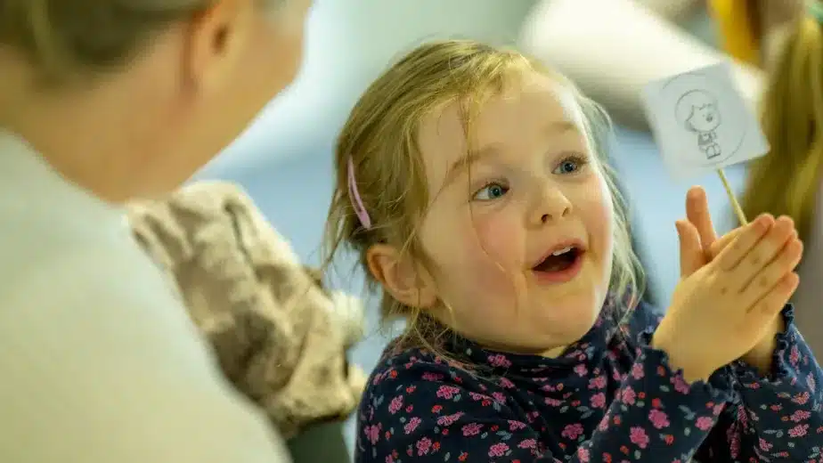 A young girl with blonde hair, wearing a floral shirt, smiles excitedly whilst holding a small stick with a drawing on it, as an adult beside her looks on.