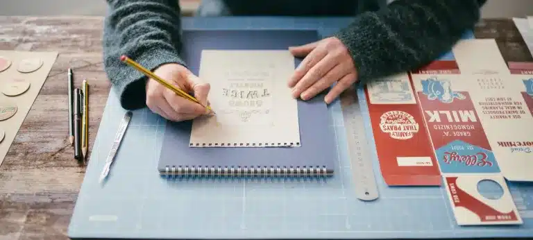 A person sketches on a notepad at a desk with a ruler, pens, and vintage-style food packaging designs on a blue cutting mat.