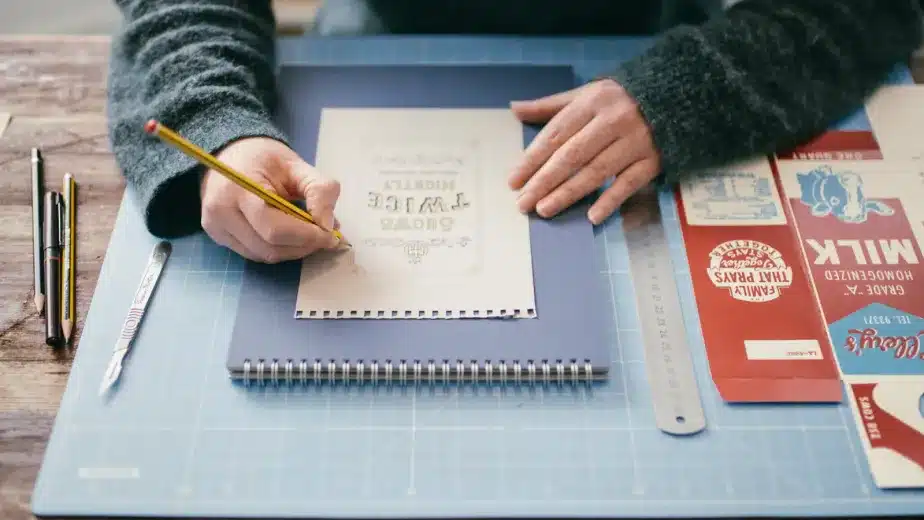 A person sketches on a notepad at a desk with a ruler, pens, and vintage-style food packaging designs on a blue cutting mat.