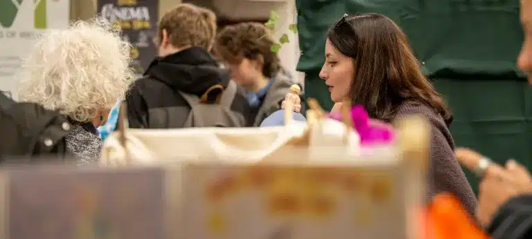 A woman with dark hair speaks to someone at a busy indoor event. Other people with rucksacks are visible in the background, and a green curtain or partition is behind them. The setting appears lively and social.
