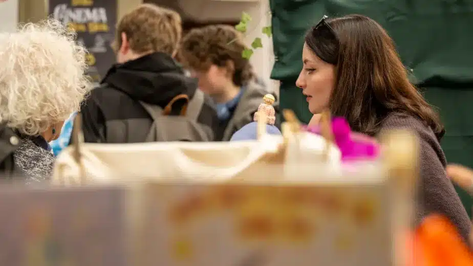 A woman with dark hair speaks to someone at a busy indoor event. Other people with rucksacks are visible in the background, and a green curtain or partition is behind them. The setting appears lively and social.