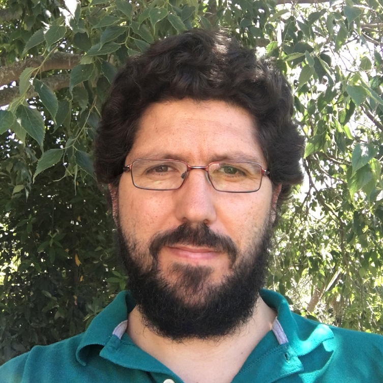 A man with glasses and a beard stands outdoors in front of leafy green trees, wearing a teal collared shirt and looking directly at the camera. Sunlight filters through the branches above him.