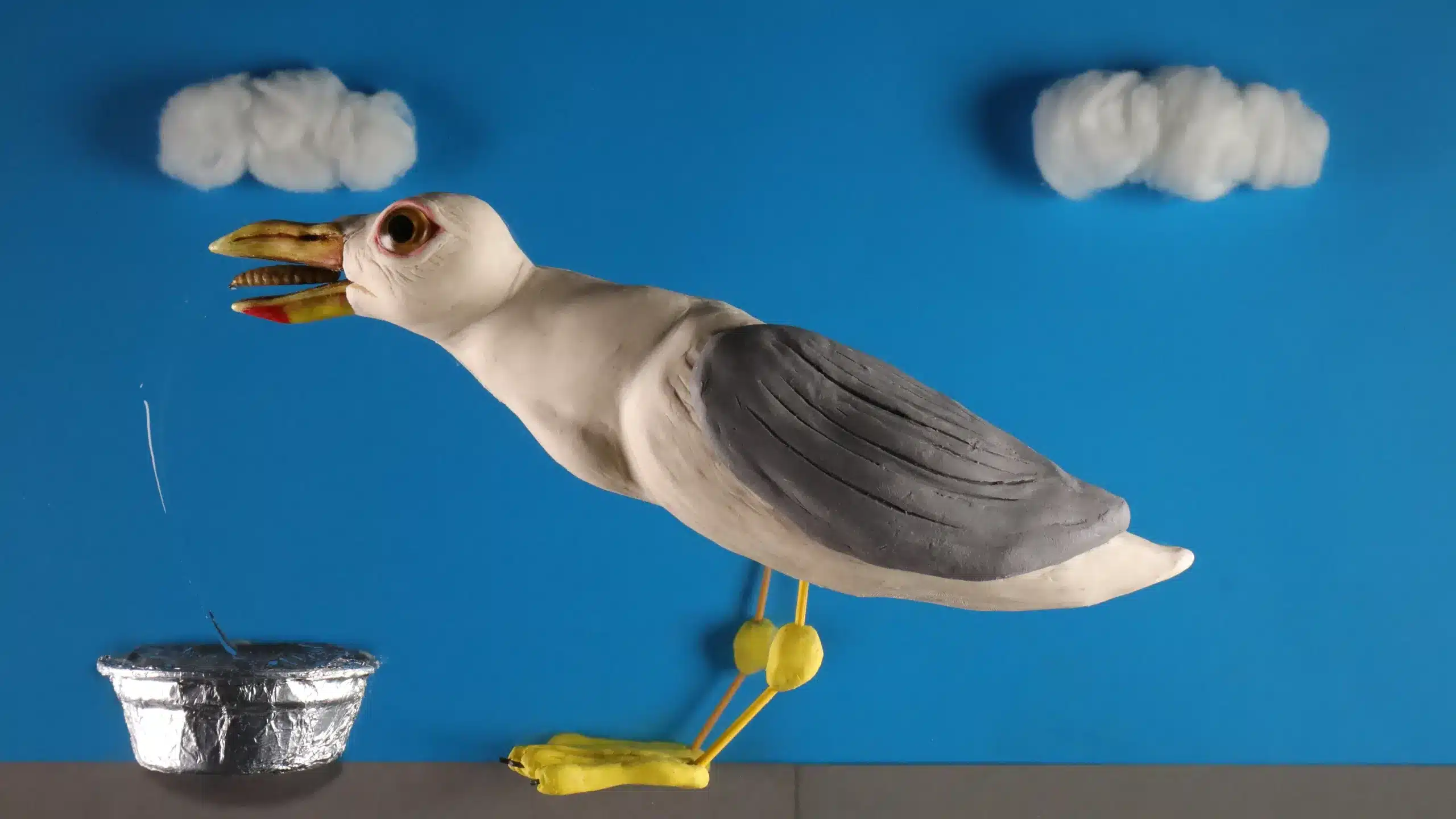A clay model of a seagull stands on a surface, reaching its beak towards a foil container. Two cotton clouds hang against a blue background, resembling a sky.